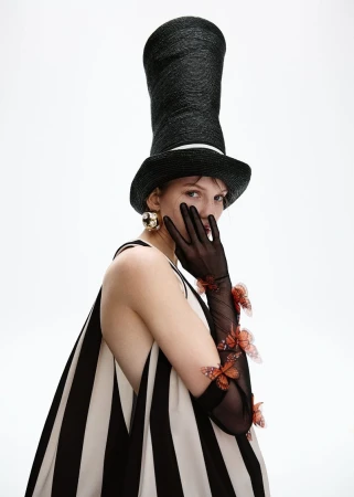 Adult woman poses in a black-and-white striped dress, a black top hat, black gloves with orange butterfly appliques against a white backdrop.