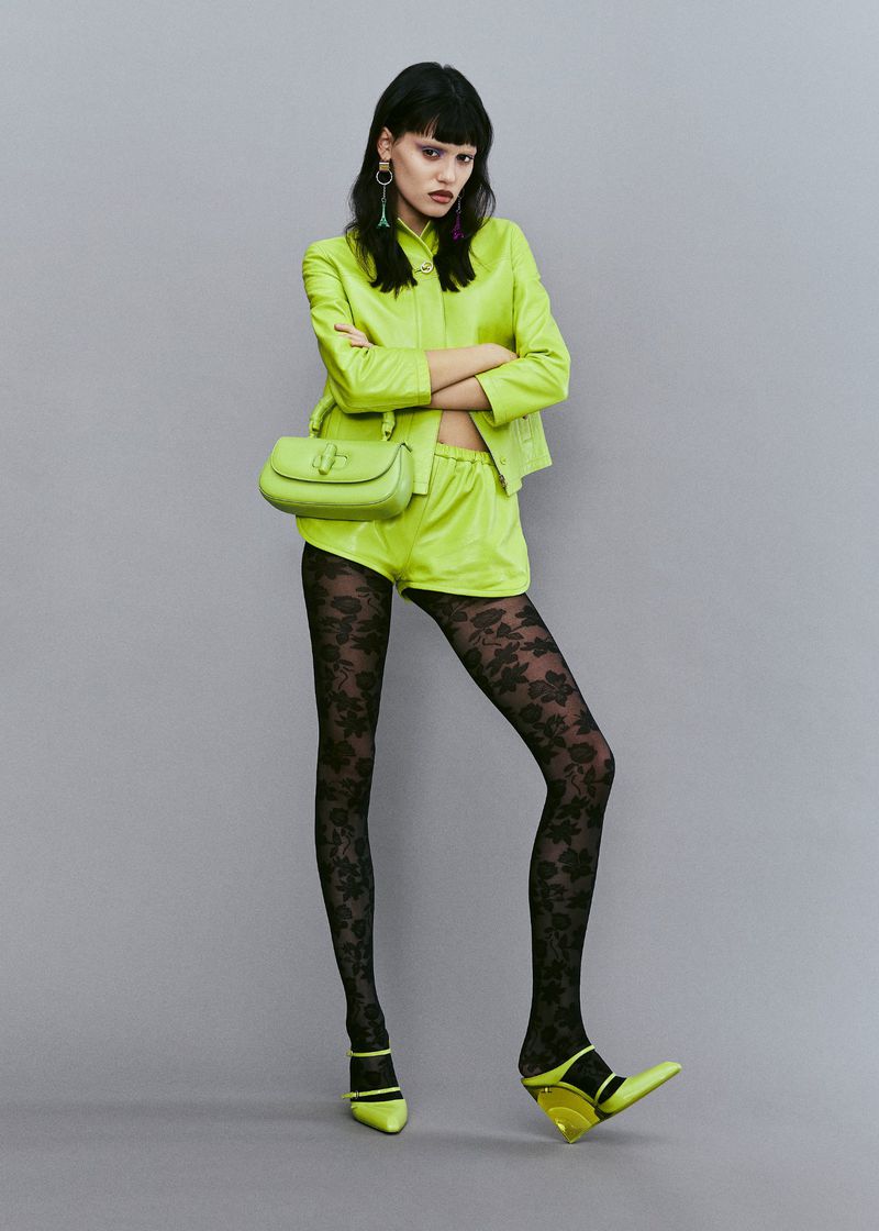 A woman in a neon green suit poses against a neutral grey background, wearing black patterned tights, matching shoes, and a purse.