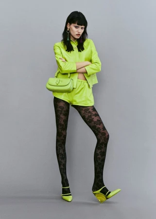 A woman in a neon green suit poses against a neutral grey background, wearing black patterned tights, matching shoes, and a purse.
