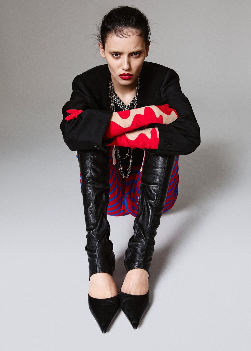 Model seated wearing a black blazer, patterned trousers, red patterned gloves, and black leather boots against a grey background.