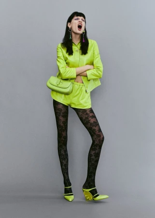 A woman in a chartreuse outfit with floral tights poses against a gray background with arms crossed and mouth open in a studio shot.