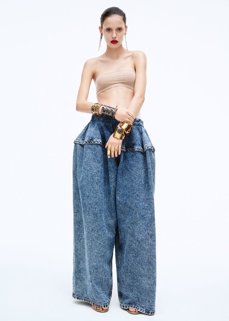 A woman poses in wide leg, light wash jeans, with a beige strapless top and large gold jewelry against a stark white backdrop.