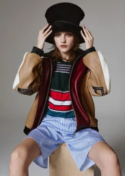 Model wearing a black top hat, color-blocked bomber jacket, and striped shorts sitting in a studio