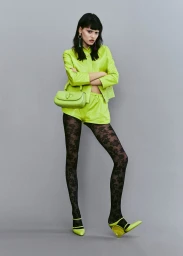 Model in neon green jacket and shorts wearing floral tights holding a green handbag against a grey backdrop