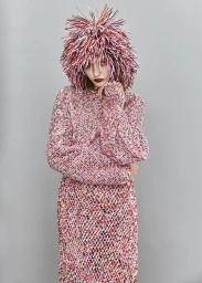 Model wearing a textured pink multi-colored fringed sweater and matching fringed headwear against a grey backdrop.