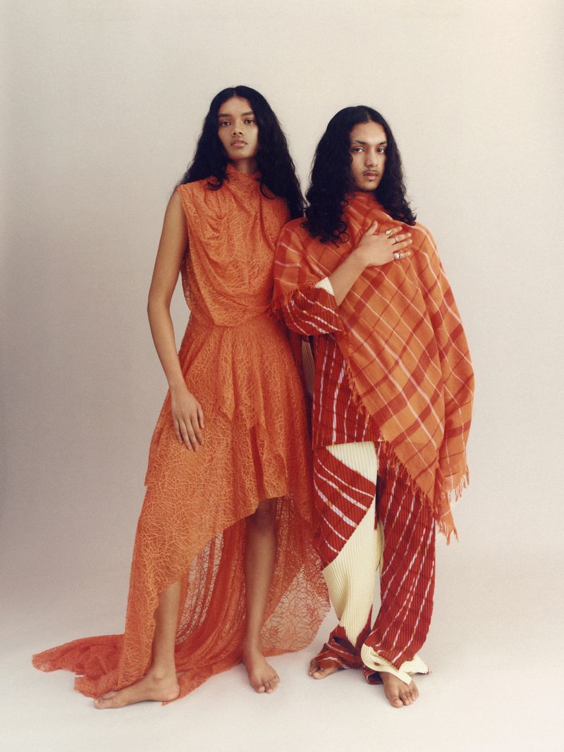 Two women in an indoor fashion editorial shot wearing orange patterned dresses and posing against a neutral backdrop.