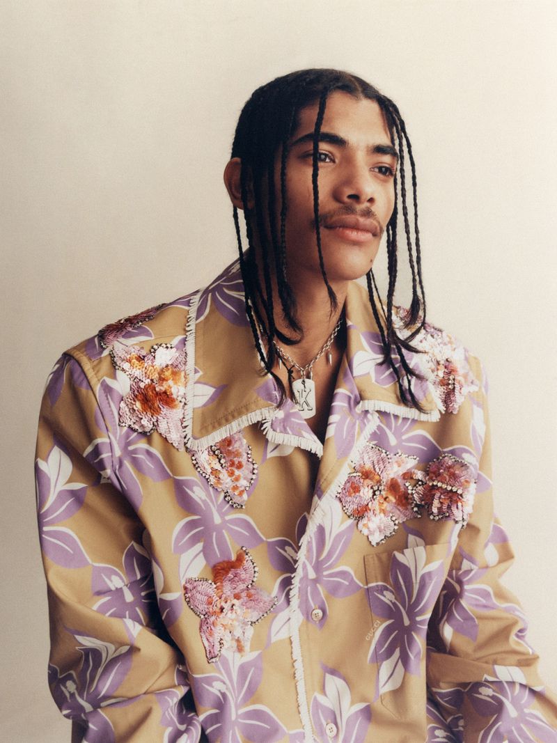 Portrait of a young adult male with braided hair wearing a tan and purple floral print shirt against a neutral studio backdrop.