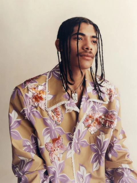 Portrait of a young adult male with braided hair wearing a tan and purple floral print shirt against a neutral studio backdrop.