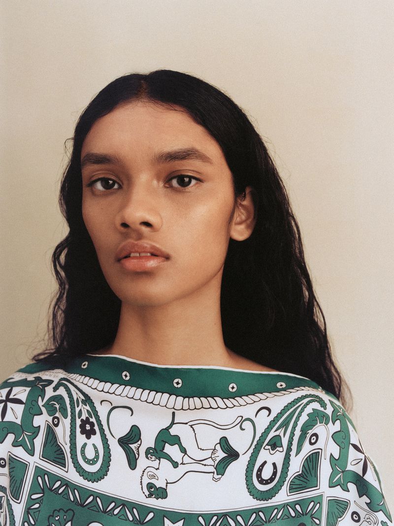 Close-up studio portrait of a young woman with long dark hair wearing a patterned top, with fair skin and neutral expression.