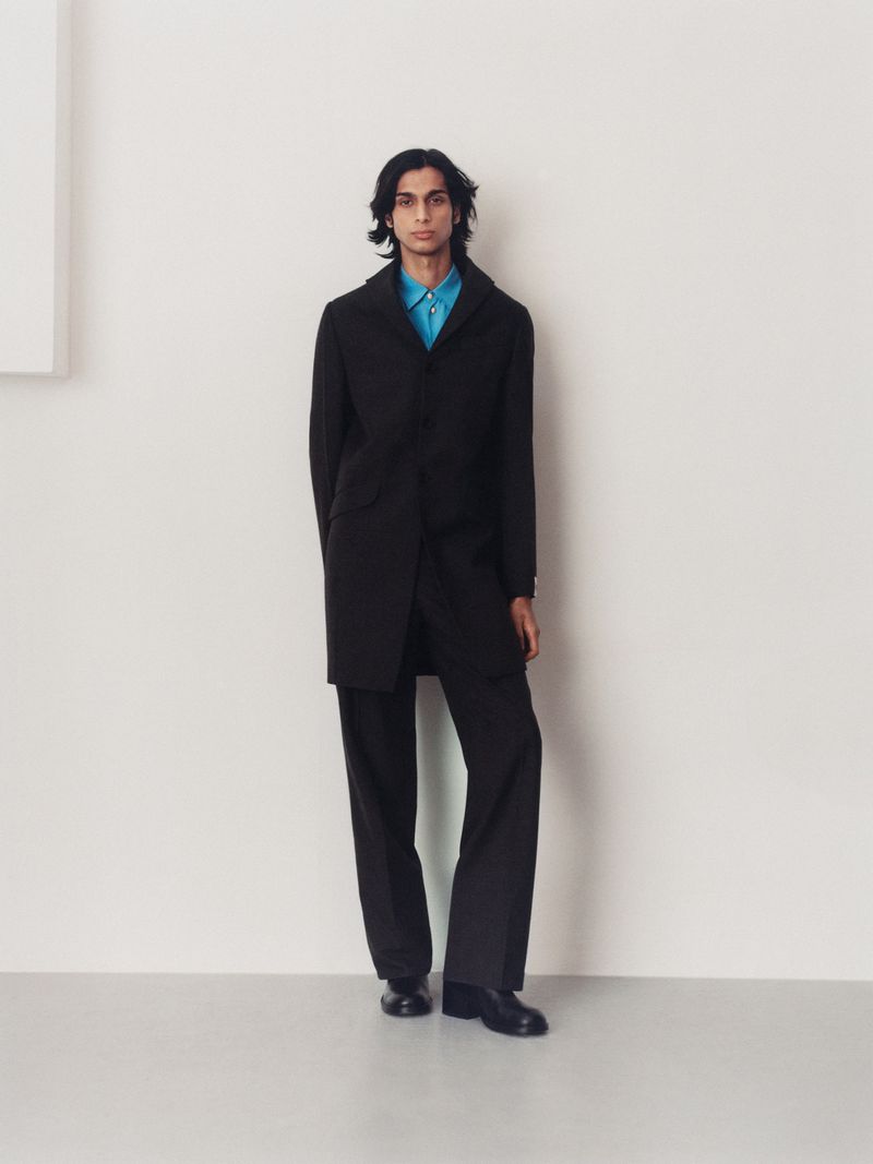 A full-length portrait of a male model in a black coat, blue shirt, and black trousers against a neutral background in a fashion editorial.
