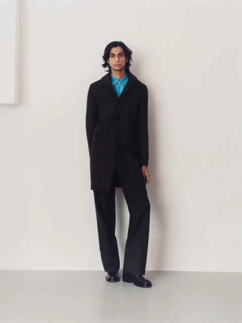 A full-length portrait of a male model in a black coat, blue shirt, and black trousers against a neutral background in a fashion editorial.