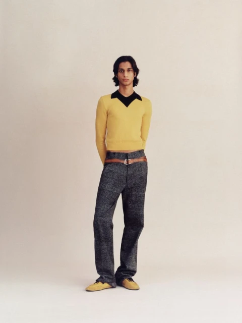 Full-length shot of a slender adult male with dark hair, wearing a yellow sweater, textured grey trousers with belt, and matching shoes.