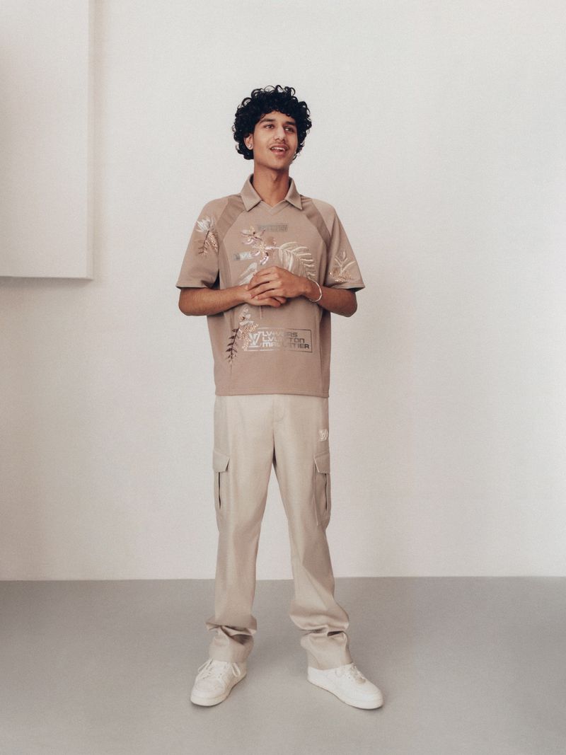 A young adult male standing in front of a white wall wearing a tan shirt with embroidery and light pants in a neutral setting.
