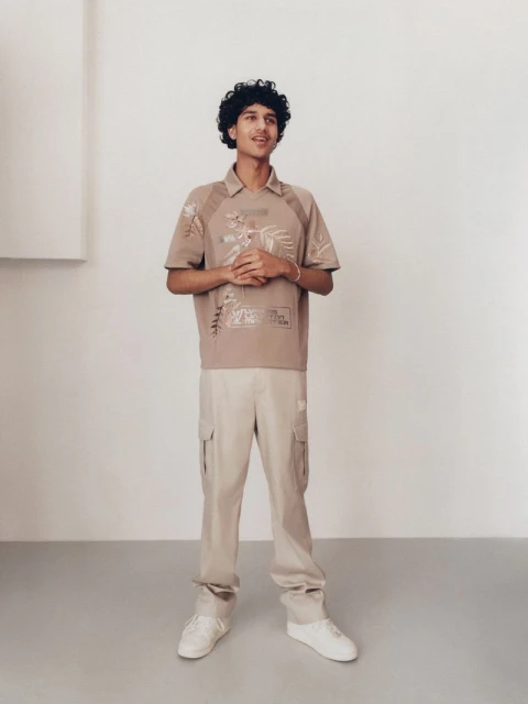 A young adult male standing in front of a white wall wearing a tan shirt with embroidery and light pants in a neutral setting.