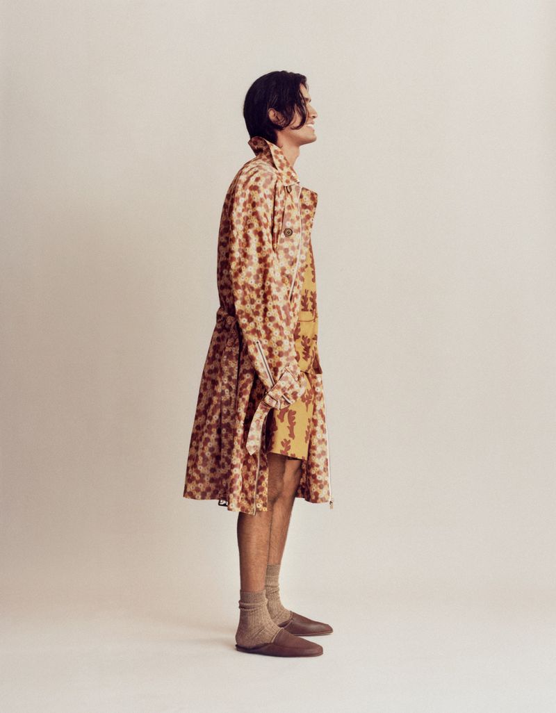 A man poses in a printed coat and shorts against a neutral backdrop. The profile shot highlights his casual yet fashionable attire.