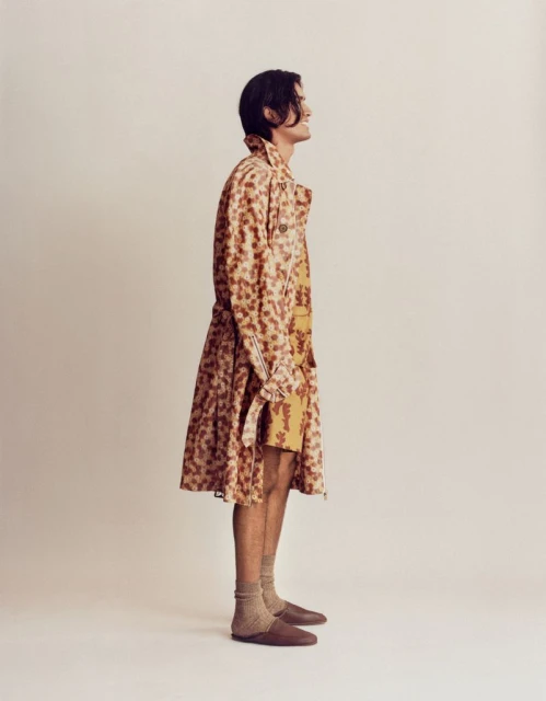 A man poses in a printed coat and shorts against a neutral backdrop. The profile shot highlights his casual yet fashionable attire.