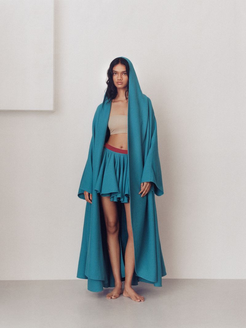 Woman poses indoors wearing a hooded teal coat and matching skirt. The background is a minimalist cream tone with a modern geometric shape.