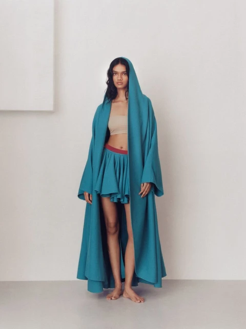 Woman poses indoors wearing a hooded teal coat and matching skirt. The background is a minimalist cream tone with a modern geometric shape.