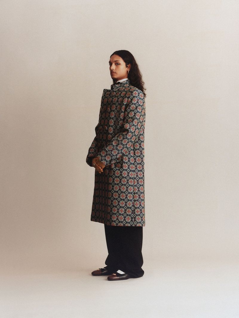 A stylish individual in a patterned coat and black pants stands against a neutral backdrop, captured in a full shot in a photo studio.