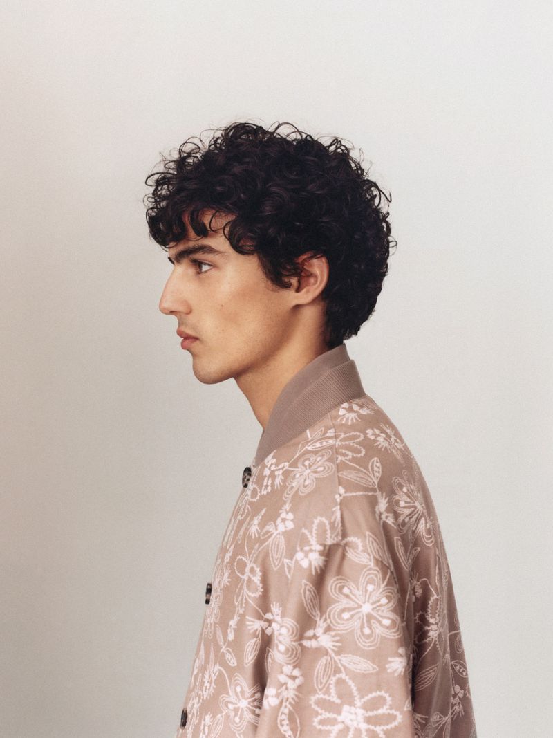 Profile of a young adult male with curly hair, wearing a light brown floral-patterned jacket against a neutral background in a studio setting.