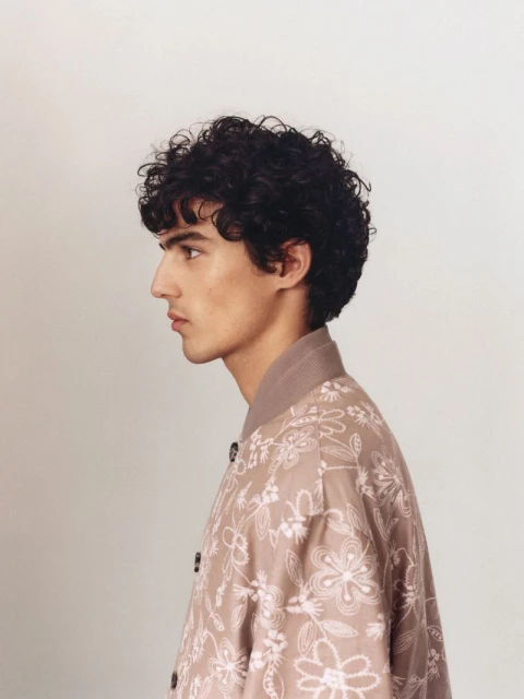 Profile of a young adult male with curly hair, wearing a light brown floral-patterned jacket against a neutral background in a studio setting.