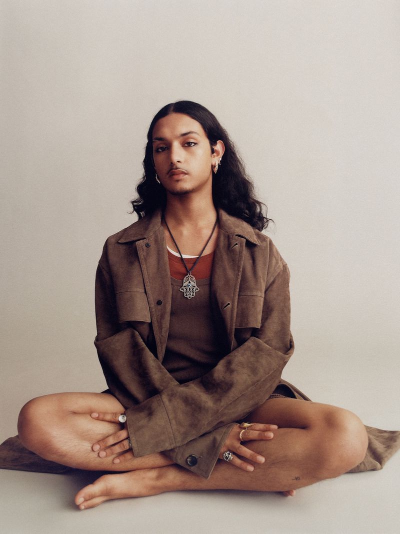 Person with long dark hair sits cross-legged in a photo studio wearing a brown jacket, dark tank top, and jewelry. Studio shot, neutral background.