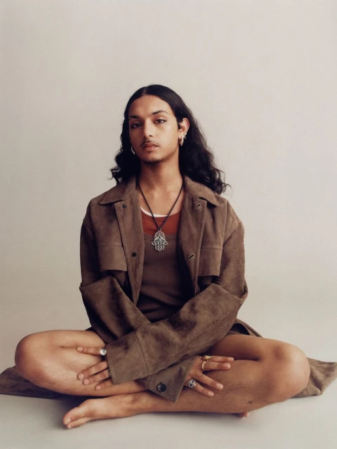 Person with long dark hair sits cross-legged in a photo studio wearing a brown jacket, dark tank top, and jewelry. Studio shot, neutral background.