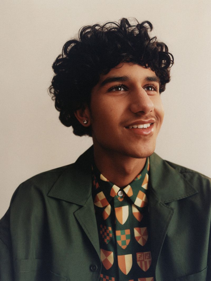 Young man with dark curly hair wearing a green jacket and patterned shirt against a plain background