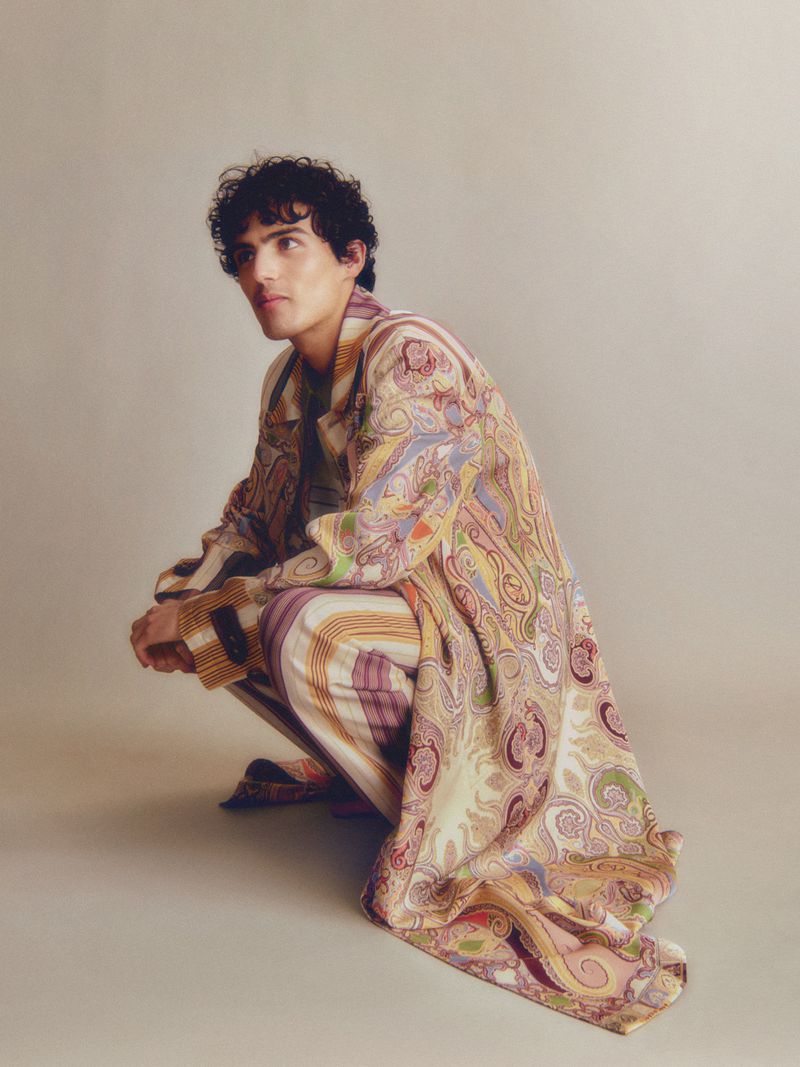 Man in paisley print coat and striped pants squats in a photo studio, looking away in a fashion pose against a neutral backdrop.