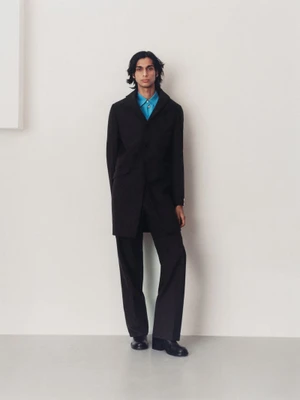 Model wearing a black coat and blue shirt standing against a plain white studio wall for The Greatest Magazine
