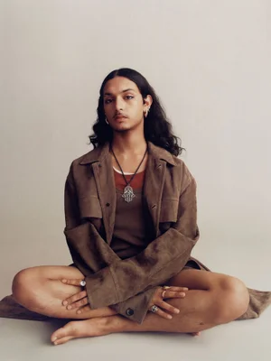 Model with long dark hair sitting cross-legged wearing a brown suede jacket over a brown top against a neutral studio backdrop