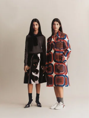 Two female models standing against a neutral studio backdrop wearing patterned and textured designer fashion apparel