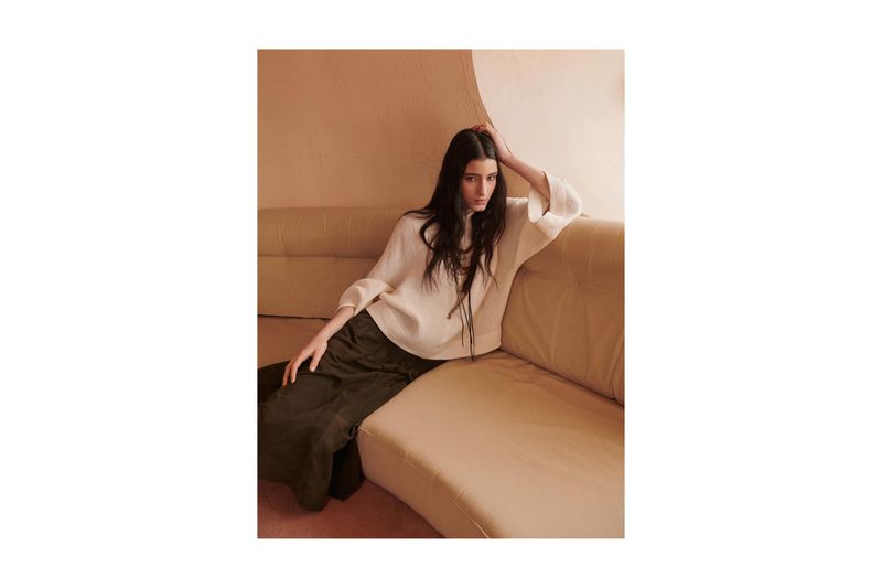 A brunette woman with long hair poses on a caramel-colored sofa wearing an off-white top and olive skirt in a studio setting.