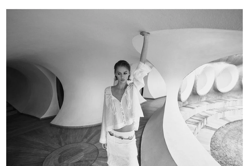 A young adult woman poses in a bright interior wearing a white blouse and skirt, with light bouncing off the curved architectural forms.
