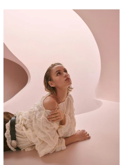 Woman in a cream dress lies on a pink surface, looking up toward a white curved structure in a fashion photography setting.