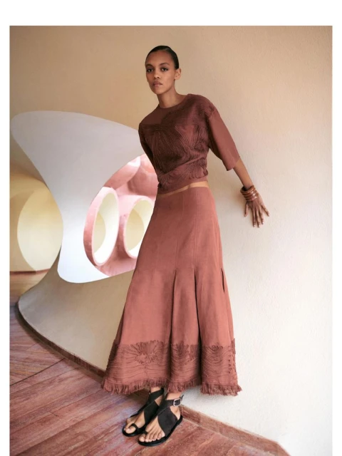 Elegant adult female poses indoors wearing a brown blouse, long brown skirt, and black strappy sandals in a fashion editorial.