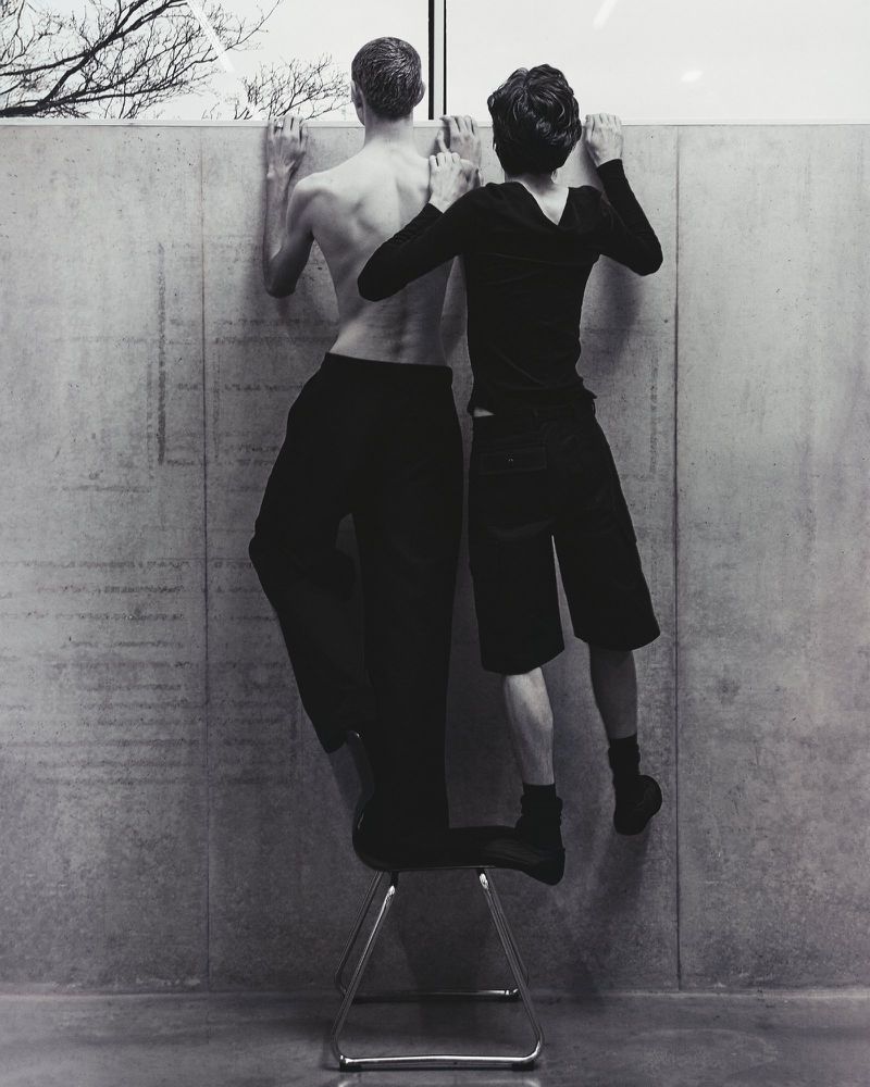 Black and white photograph of two people standing on a chair against a concrete wall, looking out a window with bare tree branches.