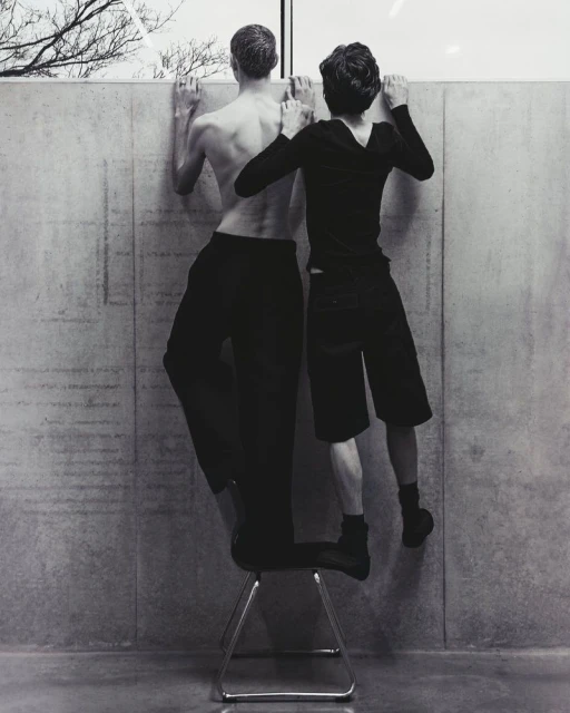 Black and white photograph of two people standing on a chair against a concrete wall, looking out a window with bare tree branches.