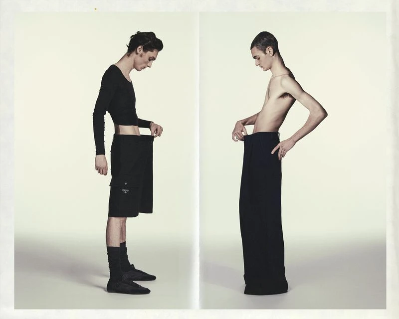 Two slim adult males posing in a studio. One wears a black long-sleeve crop top and skirt, while the other is shirtless in long black pants.