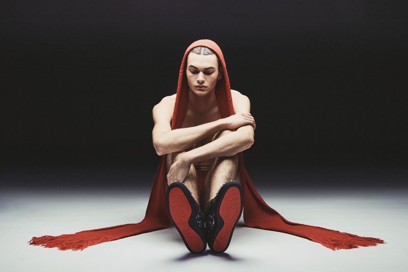 A pale-skinned adult male is seated on the floor with a long red knit scarf wrapped around his head and shoulders against a dark background.