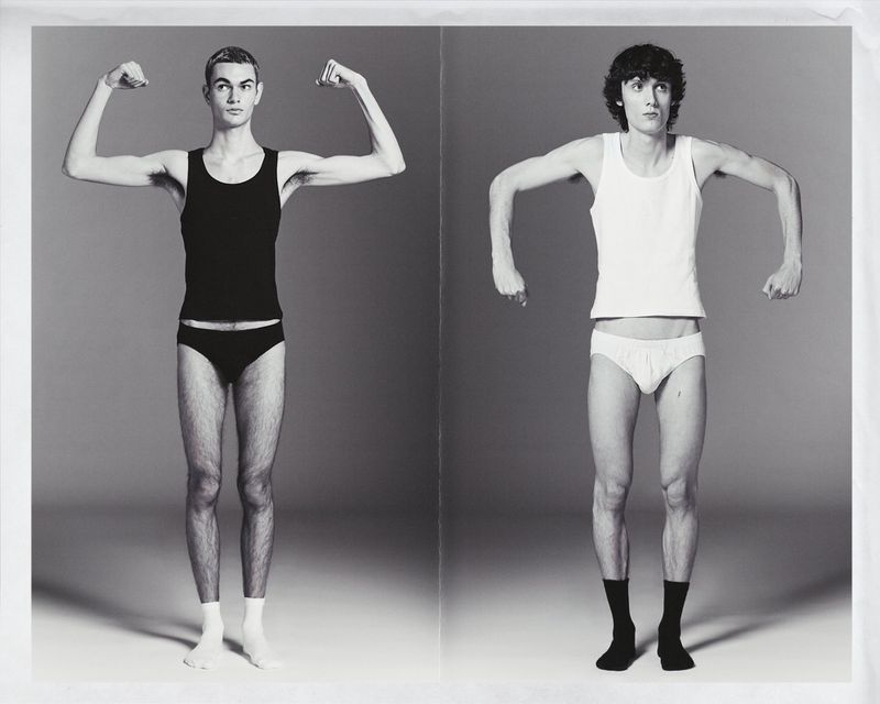 Two thin men stand in underwear and socks, flexing biceps in a black and white photo studio shot against a seamless gray background.