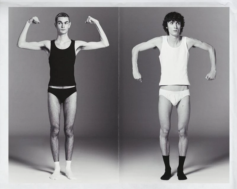 Two thin men stand in underwear and socks, flexing biceps in a black and white photo studio shot against a seamless gray background.