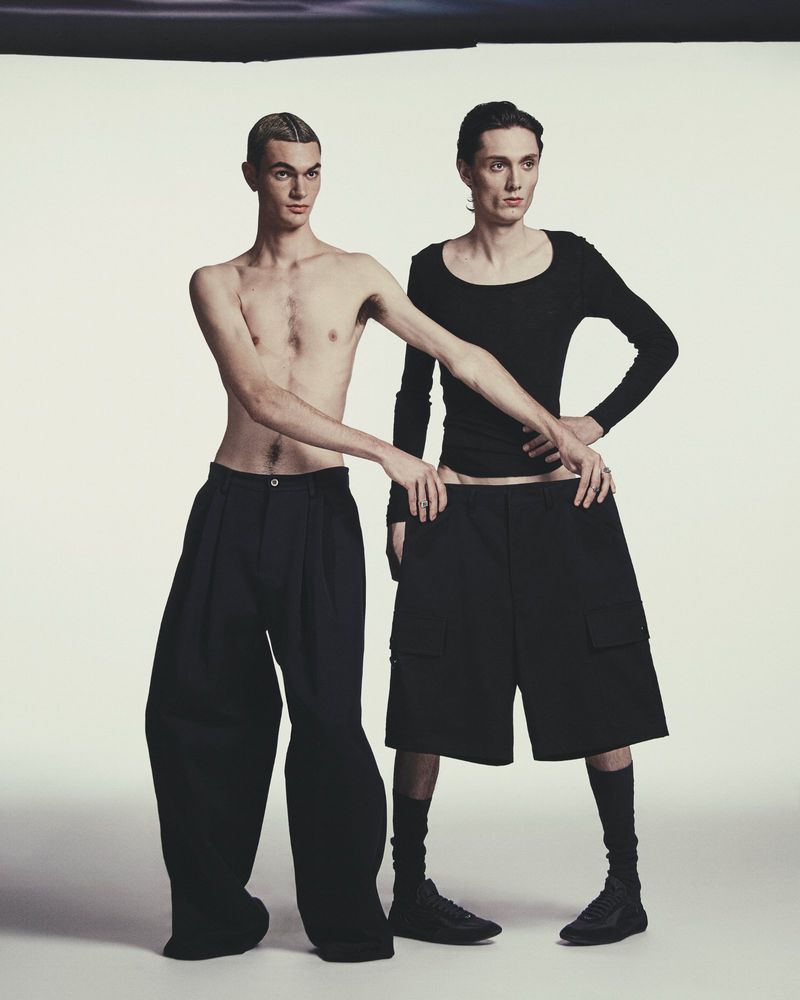 Two male models posing together against a plain white studio backdrop, wearing oversized black trousers and dark footwear.