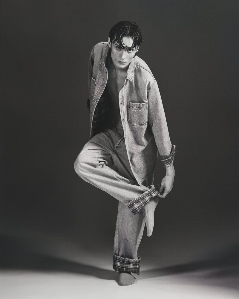 Black-and-white portrait of a young adult male fashion model posing with bare feet in a denim shirt and jeans with plaid cuffs.