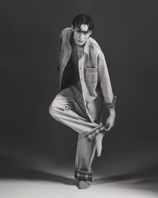 Black-and-white portrait of a young adult male fashion model posing with bare feet in a denim shirt and jeans with plaid cuffs.
