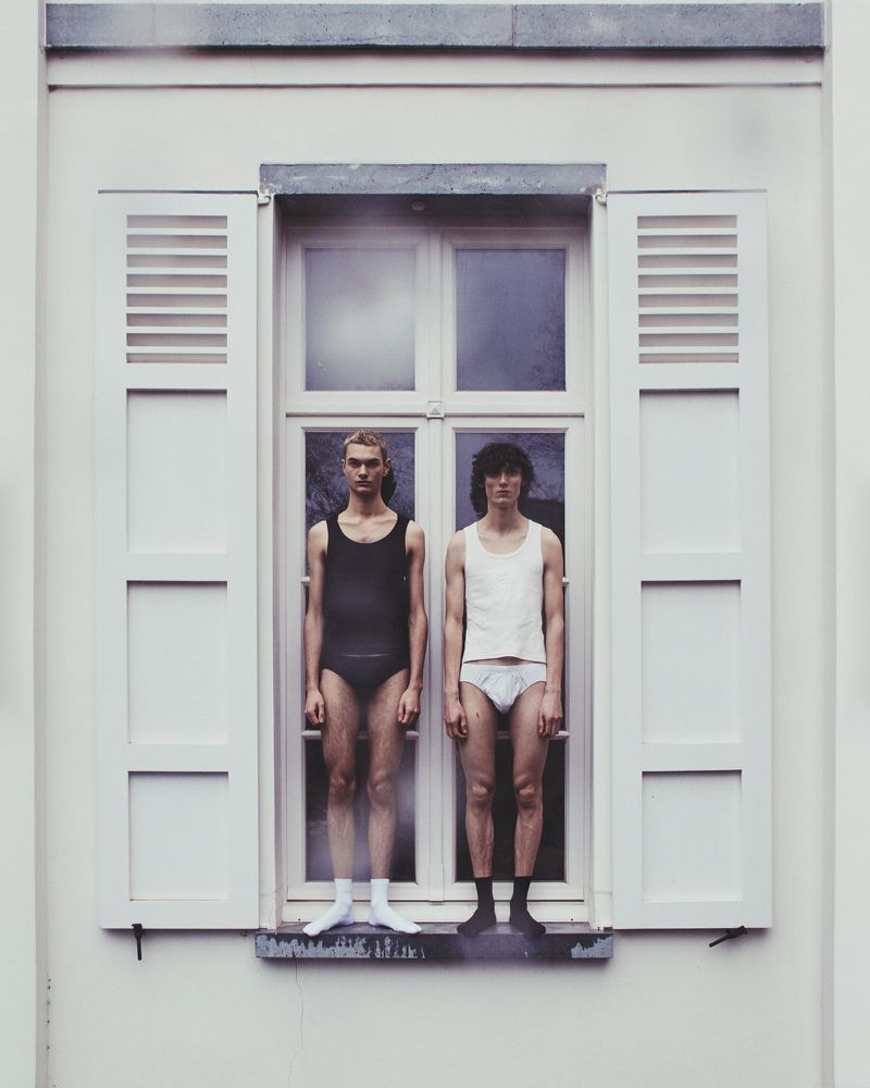 Two fair-skinned adult men in minimal attire stand side-by-side in a window with white shutters, creating a stark, symmetrical composition.