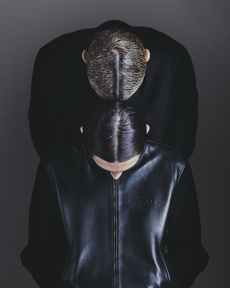 Fashion shot featuring two people with slicked back hair and dark clothing against a grey background in a studio setting. Overhead shot.