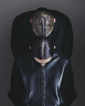 Two models with slicked-back hair wearing black jackets from above against a dark grey seamless studio background.