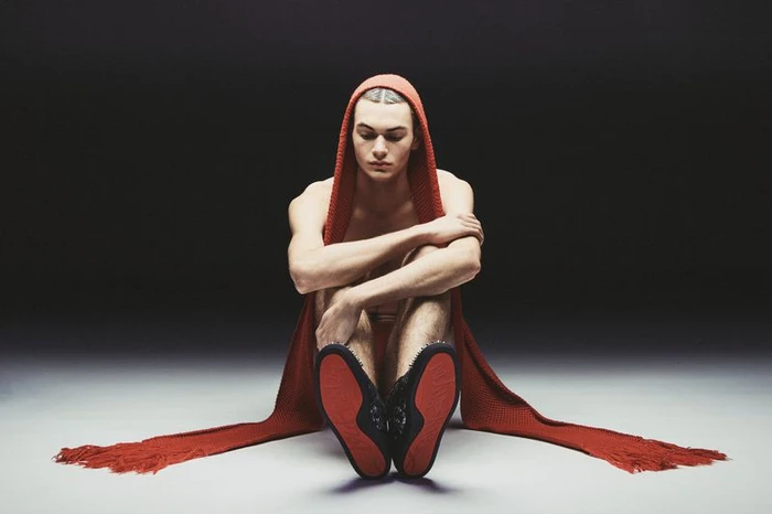 A young male model sits on a grey floor wearing a red hooded scarf and dark shoes with red soles, photographed by Sieme Hermans.
