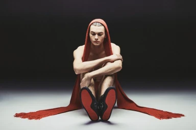 A young male model sits on a grey floor wearing a red hooded scarf and dark shoes with red soles, photographed by Sieme Hermans.